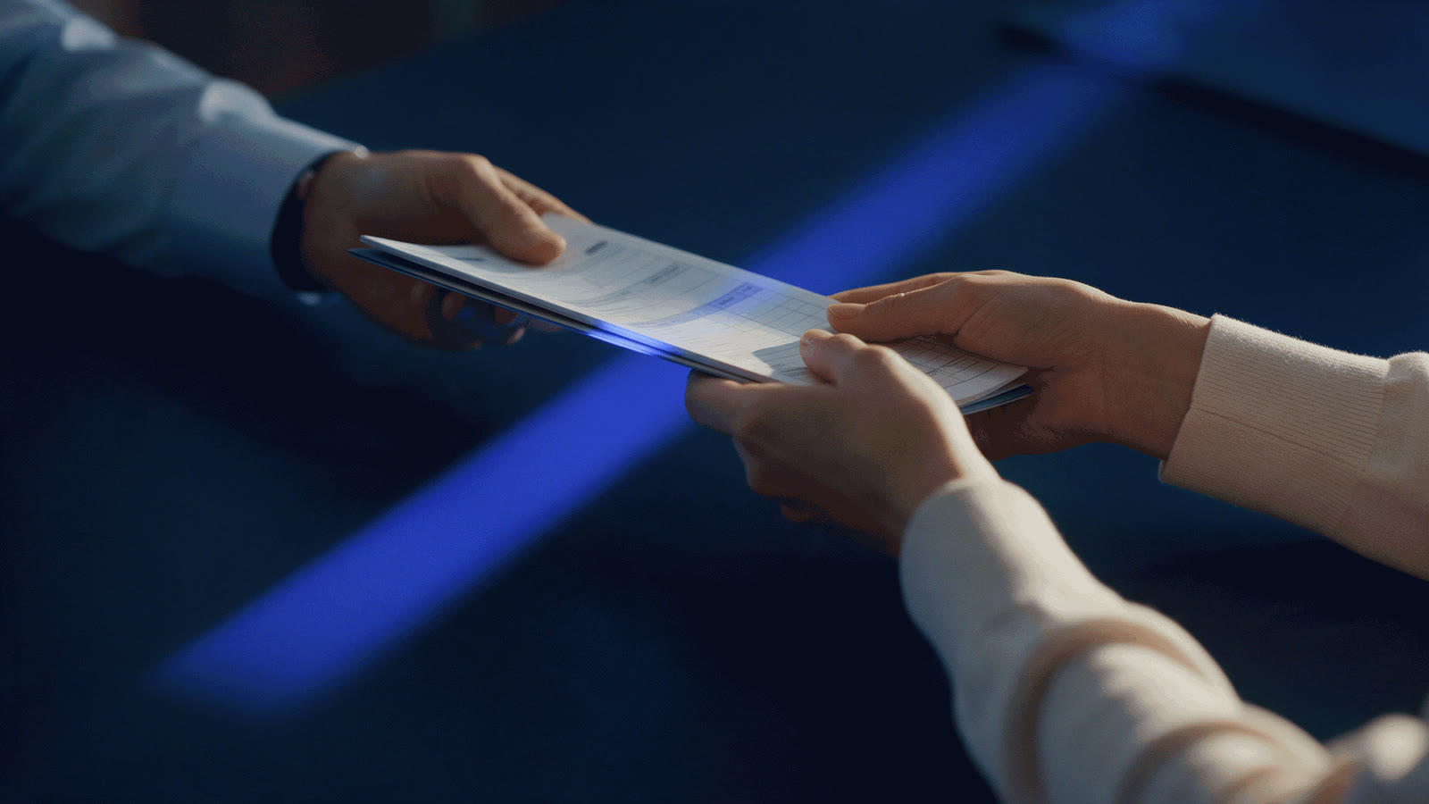 One person handing a set of claim forms to another across a navy desk, crossed by a band of blue light